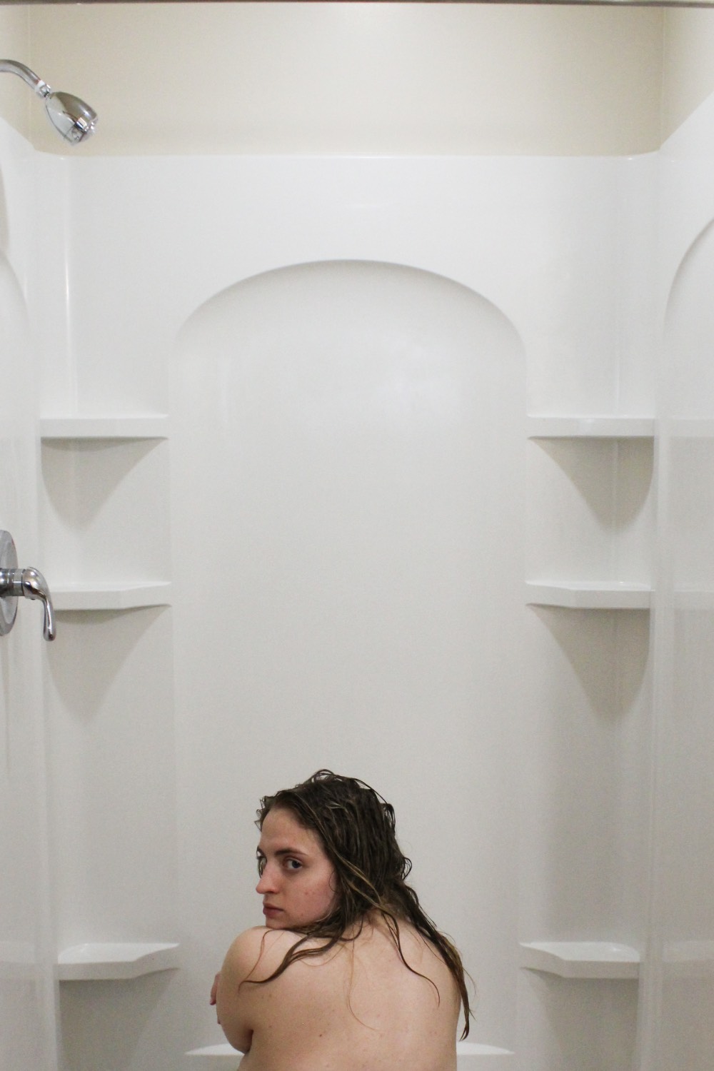 Color photo of a young woman sitting in a shower looking over her shoulder back at the camera